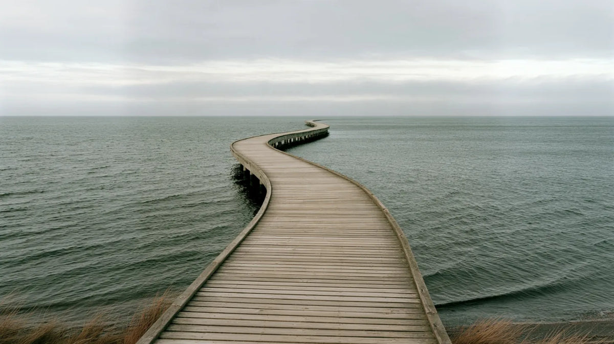 Langer Holzsteg, der in sanften Kurven weit ins ruhige Meer hinausführt, unter bedecktem Himmel.