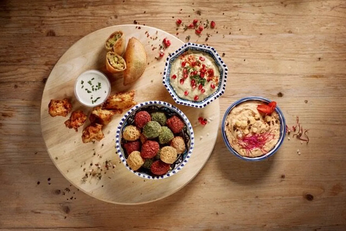 Falafel in Rot, Grün und Beige, zwei Schalen Hummus, Samosas und ein heller Dip auf einem runden Holzbrett – Vegetarian Quality
